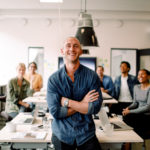 Portrait of smiling male entrepreneur standing with arms crossed and employees sitting at office desk