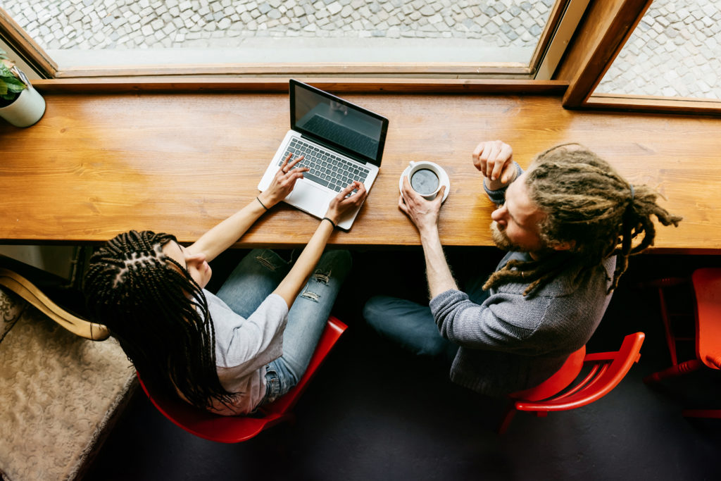 A young man and a young woman are talking to each other in a bright café. Both of them have dark dreadlocks. She's working on a notebook while he is drinking some coffee.